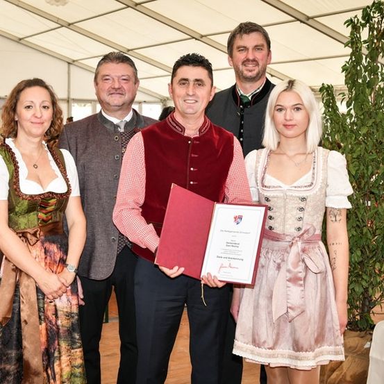 Fünf Personen stehen lächelnd und posieren für ein Foto in einem Raum. Ein Mann hält ein Zertifikat und eine Mappe. Hinter ihnen sind eine Pflanze und ein Tisch mit einer weißen Tischdecke zu sehen.