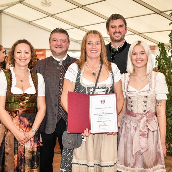 Fünf Menschen stehen lächelnd in einem Zelt für ein Foto. Die Frau in der Mitte hält ein Zertifikat. Hinter ihnen stehen und plaudern weitere Menschen.