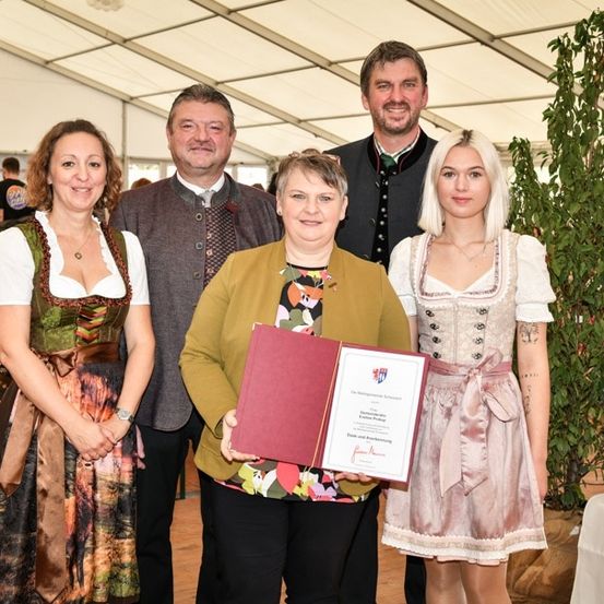 Eine Gruppe von Menschen steht in einem Zelt und lächelt für ein Foto. Eine Frau hält ein Zertifikat, und zwei Frauen in traditionellen Kleidern stehen neben ihr. Ein Mann in einem Anzug steht hinter ihnen.