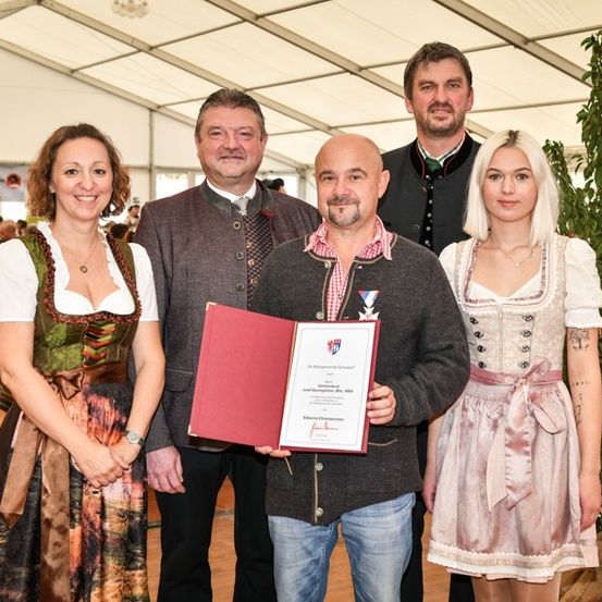 Eine Gruppe von Menschen in traditioneller Kleidung, die lächelnd und mit einem Mann, der ein Zertifikat hält, für ein Foto posieren. Dahinter ein Zelt mit einer Menschenmenge.