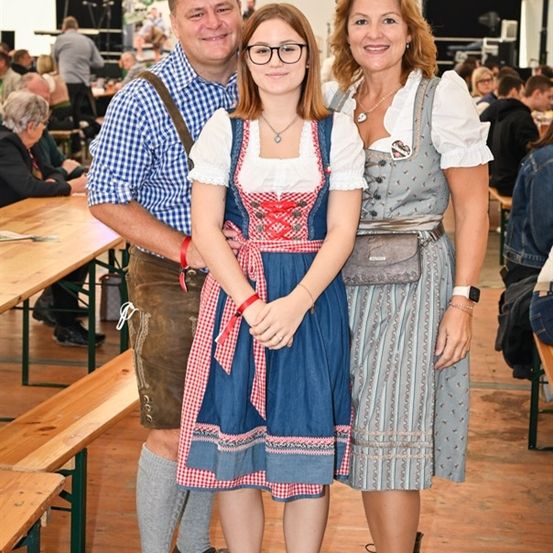 Eine Familie aus drei Personen, gekleidet in traditioneller Kleidung, steht lächelnd für ein Foto in einer festlichen Umgebung. Der Mann trägt ein kariertes Hemd und Hosenträger, die Frau ein traditionelles Kleid und das Mädchen ein passendes Outfit. Sie sind von einem verblassten Hintergrund von Menschen bei einem Treffen umgeben.