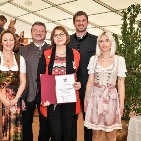 Fünf Menschen stehen lächelnd unter einem Zelt. Eine Frau in der Mitte hält ein Zertifikat. Hinter ihnen ist eine große Topfpflanze und ein Tisch mit einem Buch zu sehen.