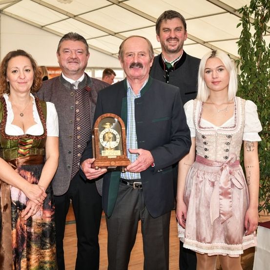 Fünf Personen stehen lächelnd und posieren für ein Foto. Der Mann in der Mitte hält eine Trophäe. Die Frauen auf beiden Seiten tragen traditionelle Kleider. Dahinter ist eine große Pflanze und eine weiße Tischdecke zu sehen.