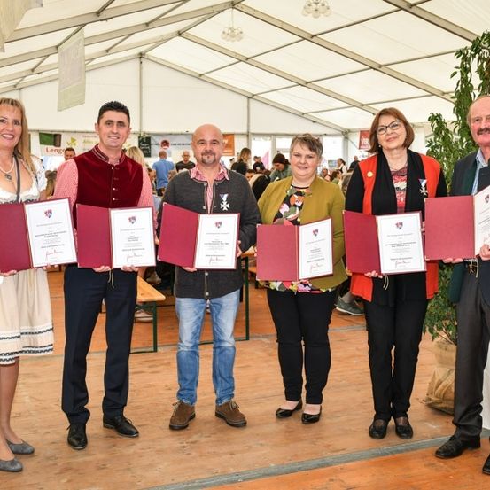 Eine Gruppe von sechs Personen steht in einem Zelt, hält Urkunden in den Händen und lächelt für ein Foto. Sie sind in verschiedenen traditionellen und formellen Kleidungsstücken gekleidet. Hinter ihnen ist eine große Pflanze und ein weißer Tisch zu sehen.