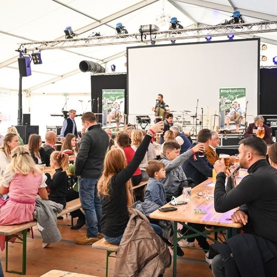 Eine Gruppe von Menschen ist in einem Zelt für eine Veranstaltung versammelt, einige sitzen an Tischen, während andere stehen und eine Aufführung auf der Bühne beobachten.