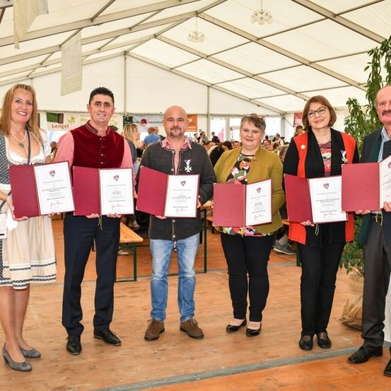Fünf Personen stehen in einer Reihe, halten rote Ordner und lächeln für ein Foto in einem Zelt mit Holzboden und weißer Decke. Hinter ihnen sind weitere Personen und eine Pflanze zu sehen.