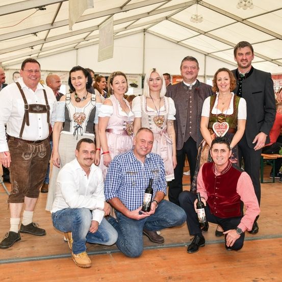 Eine Gruppe von Menschen in traditioneller bayerischer Kleidung posiert für ein Foto in einem Zelt, einige halten Flaschen Wein.