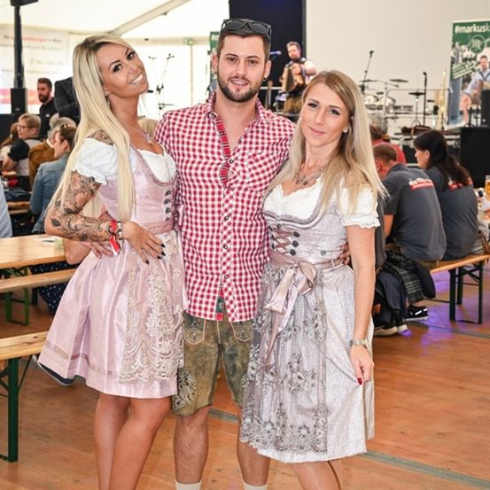 Zwei Frauen in traditioneller deutscher Kleidung stehen mit einem Mann in einem karierten Hemd auf einem Festival, mit einer Band im Hintergrund.