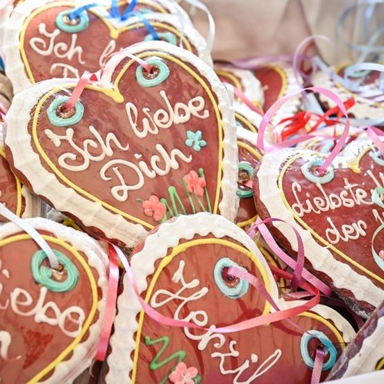 Ein Nahaufnahme von herzförmigen Keksen mit dekorativer Glasur, Bändern und deutschem Text. Die Kekse haben verschiedene Phrasen wie 'Ich liebe Dich' und 'liebste der Welt'.