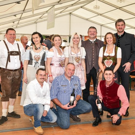Eine Gruppe von Menschen in traditioneller deutscher Kleidung, die für ein Foto in einem Zelt mit Holzböden und weißen Zelten posiert. Einige halten Bierflaschen.