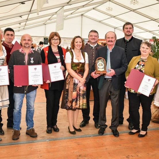 Eine Gruppe von Menschen in traditioneller Kleidung, die Urkunden und Auszeichnungen halten, steht in einem Zelt mit Holzboden und weißer Decke.