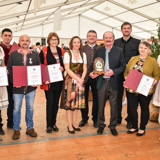 Eine Gruppe von Menschen in traditioneller Kleidung steht in einem Zelt und hält Urkunden und Auszeichnungen. Sie lächeln und posieren für ein Foto. Die Umgebung ist drinnen mit einem Holzboden und einem weißen Zelt.