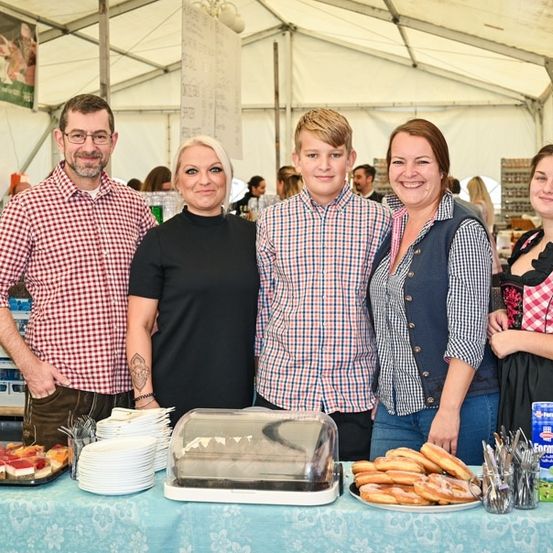 Fünf Personen stehen lächelnd hinter einem Tisch mit Essen und posieren für ein Foto. Der Tisch hat Teller, ein Brotkorb und eine Milchflasche. Im Hintergrund ist ein weißes Zelt mit einem Banner zu sehen.