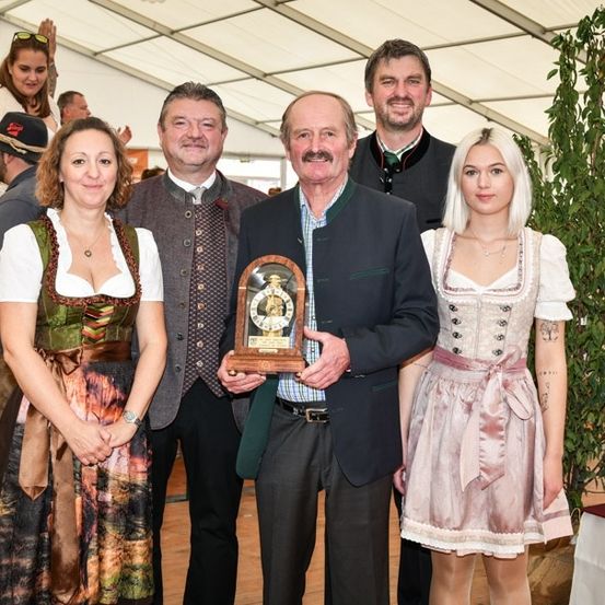 Eine Gruppe von Menschen in traditioneller Kleidung steht zusammen und hält eine Trophäe. Eine Frau links trägt ein grünes Kleid, während die Frau rechts ein rosa Kleid trägt. Ein Mann in der Mitte hält die Trophäe.