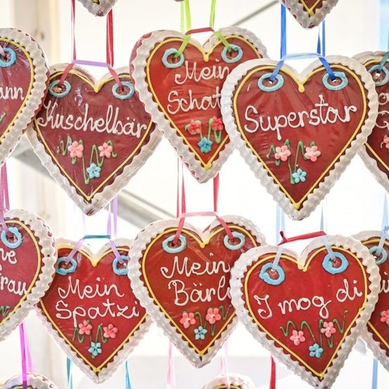 Eine Sammlung von herzförmigen Keksen mit deutschem Text darauf, wie 'Mein Schatzi' und 'Superstar', hängen an bunten Bändern. Die Kekse sind mit blauen Blumen verziert und haben eine glänzende Glasur.