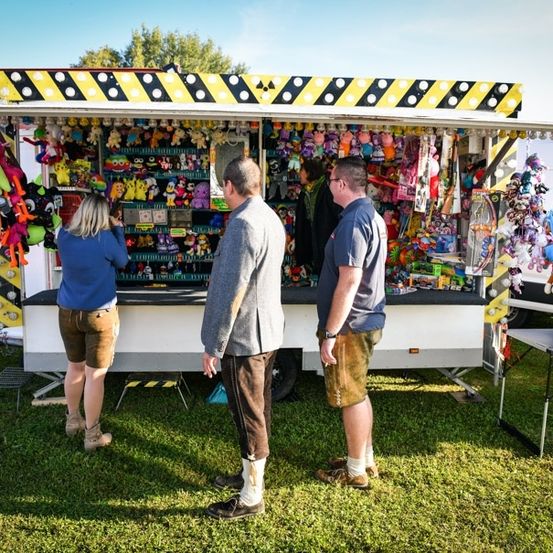 Drei Personen stehen vor einem Jahrmarktstand voller Plüschtiere und Spielzeug, wobei eine Frau die Spielzeuge betrachtet und zwei Männer einander ansehen.