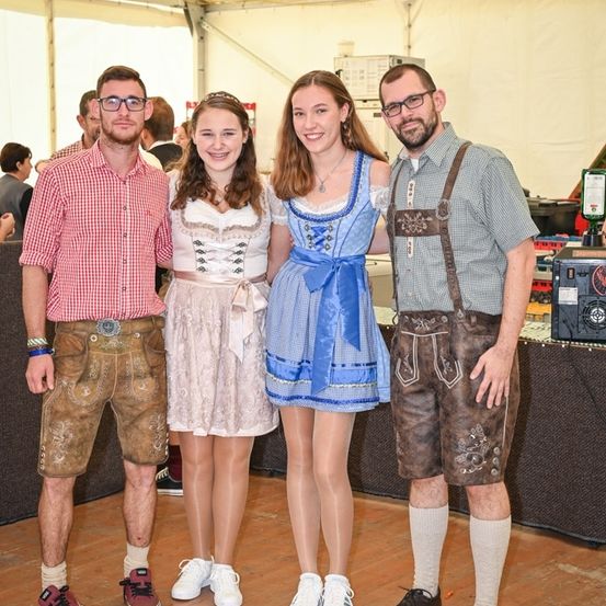 Vier Personen in traditioneller bayerischer Kleidung stehen gemeinsam in einem Raum und lächeln für die Kamera. Die beiden Männer tragen Hosenträger und Ledershorts, während die beiden Frauen traditionelle Kleider tragen.