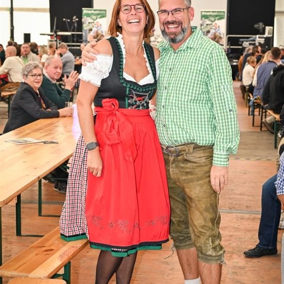 Ein Mann und eine Frau in traditioneller bayerischer Kleidung stehen in einem Biergarten, lächeln für die Kamera. Dahinter ist ein langer Holztisch aufgebaut, und mehrere Personen sitzen an Tischen.
