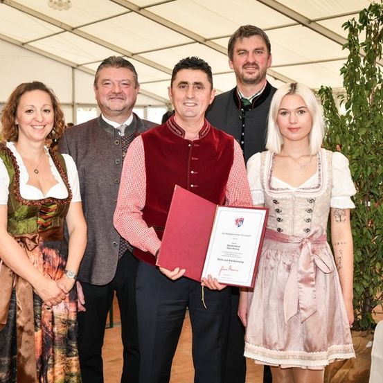 Fünf Personen stehen lächelnd in einem Zelt für ein Foto. Der Mann in der Mitte hält eine rote Mappe und ein Zertifikat. Zwei Frauen auf beiden Seiten tragen traditionelle Kleider. Hinter ihnen ist eine große grüne Pflanze und eine weiße Tischdecke zu sehen.