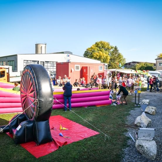 Ein farbenfrohes Outdoor-Event mit aufblasbaren Attraktionen, einer Menschenmenge und verschiedenen Ständen auf einer Rasenfläche.