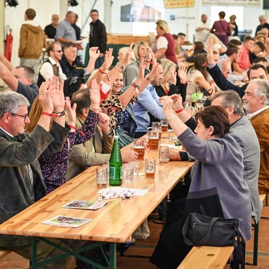 Eine große Gruppe von Menschen sitzt an einem langen Holztisch, hebt die Hände und trinkt Bier. Einige tragen Brillen, Armbänder und Uhren. Auf dem Tisch stehen Flaschen, Gläser und Papiere. Hinter ihnen sitzen und stehen weitere Menschen. Im Hintergrund befinden sich auch einige Objekte und ein Feuerlöscher.