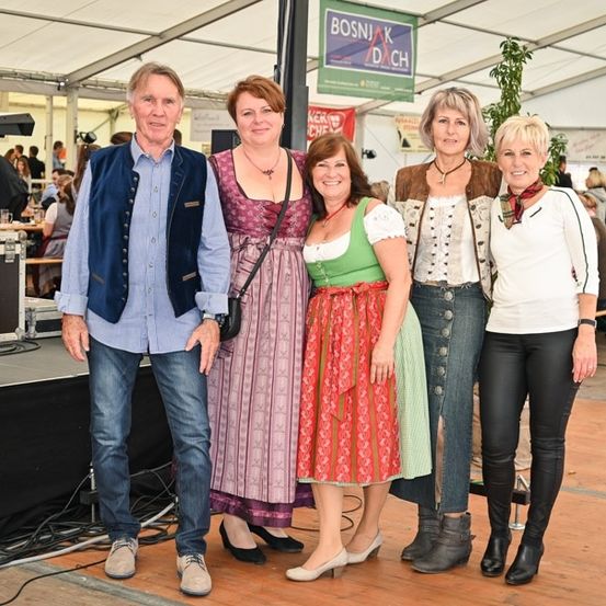 Fünf Erwachsene, drei Frauen und zwei Männer, stehen auf einer Bühne mit einem Hintergrund, auf dem 'BOSNJAK DACH' steht. Die Frauen sind in traditioneller bayerischer Kleidung gekleidet, und die Männer tragen Freizeitkleidung. Hinter ihnen sitzen Leute an Tischen, und eine Bühne mit einem Keyboard und einem Mikrofon ist sichtbar.