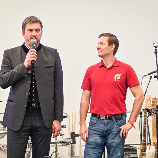Zwei Männer auf einer Bühne, einer mit einem Mikrofon und der andere mit einem roten Poloshirt, neben Musikinstrumenten und einem Banner stehend.