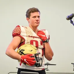 Ein Footballspieler in einem rot-weißen Trikot mit der Nummer 01 hält einen goldenen Helm und steht vor einem Mikrofon.
