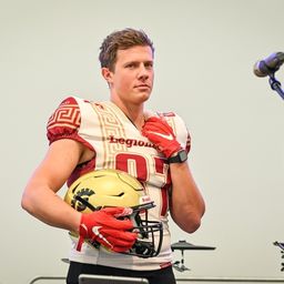 Ein Footballspieler in einem rot-weißen Trikot mit der Nummer 01 hält einen goldenen Helm und steht vor einem Mikrofon.