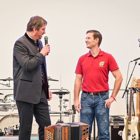 Zwei Männer auf der Bühne, einer hält ein Mikrofon und spricht, der andere in einem roten Poloshirt mit Logo, neben einem Akkordeon und verschiedenen Musikinstrumenten.