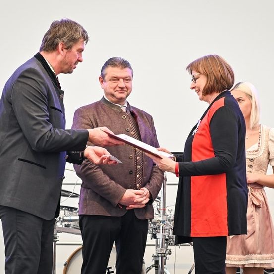 Ein Mann in einem Anzug überreicht einer Frau in einem roten Oberteil eine Urkunde, während ein Mann in einem braunen Jackett und eine Frau in einem rosa Kleid hinter ihnen stehen, in der Nähe eines Schlagzeugs und eines Mikrofons.