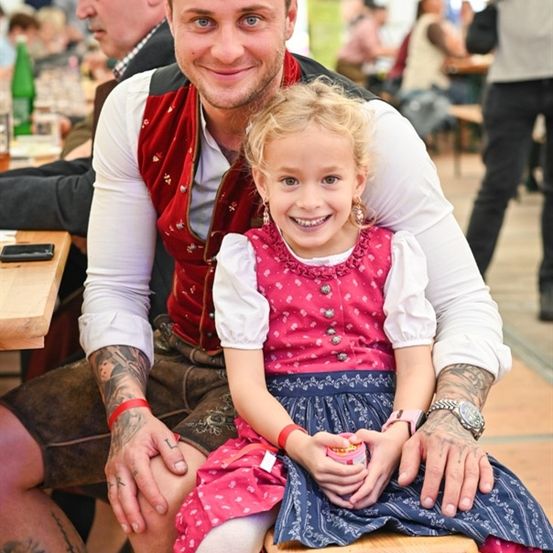 Ein Mann mit Tattoos sitzt auf einem Holzhocker mit einem lächelnden Mädchen auf seinem Schoß. Sie tragen beide traditionelle Kleidung. Hinter ihnen sitzen Menschen an Tischen und einige stehen.