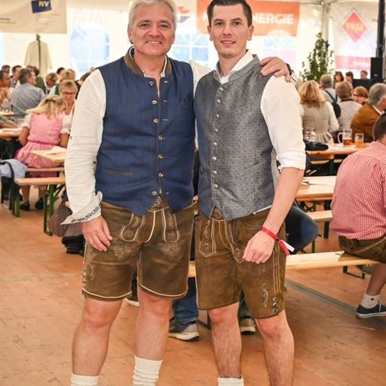 Zwei Männer in traditioneller deutscher Kleidung stehen bei einem Festival nebeneinander und lächeln für die Kamera. Dahinter sitzt eine Menge Menschen an Tischen und genießt Getränke und Essen.