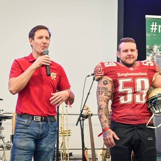 Zwei Männer stehen auf einer Bühne, einer hält ein Mikrofon und spricht, der andere hält einen Footballhelm und schaut den Sprecher an. Hinter ihnen ist ein Banner mit einem Hashtag und einem Baum.