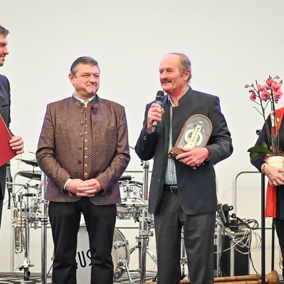 Vier Personen stehen auf einer Bühne, einer hält ein Mikrofon und eine Trophäe, ein anderer eine rote Mappe, und zwei weitere lächeln. Hinter ihnen sind Musikinstrumente und ein weißer Hintergrund zu sehen.