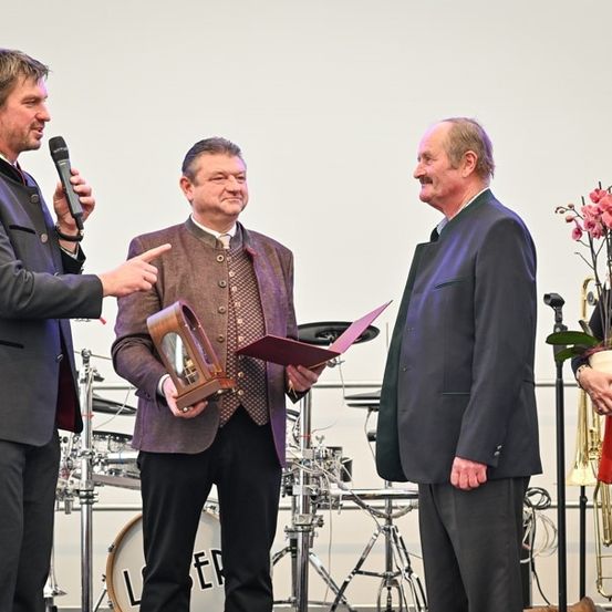Ein Mann mit Mikrofon spricht zu einem Mann, der eine Trophäe und ein Dokument hält. Hinter ihnen hält eine Frau einen Blumenstrauß. Im Hintergrund stehen Musikinstrumente.