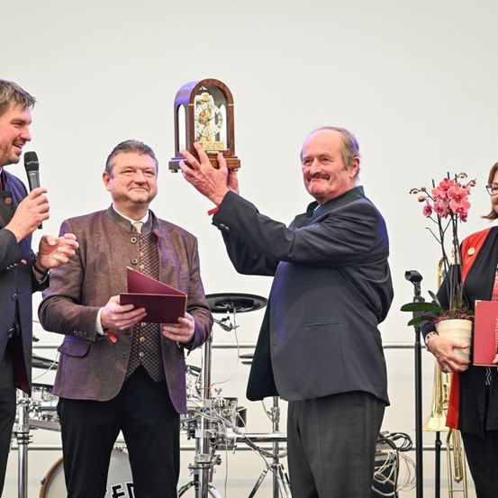 Eine Gruppe von Menschen auf einer Bühne, einer hält eine Trophäe, ein anderer ein Mikrofon und zwei weitere halten Bücher. Im Hintergrund stehen ein Schlagzeug und eine Trompete.