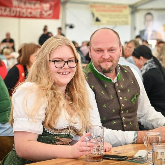 Ein Mann und eine Frau in traditioneller deutscher Kleidung sitzen an einem Tisch, lächeln und halten Biergläser. Sie sind von einer Menge auf einem Fest umgeben.