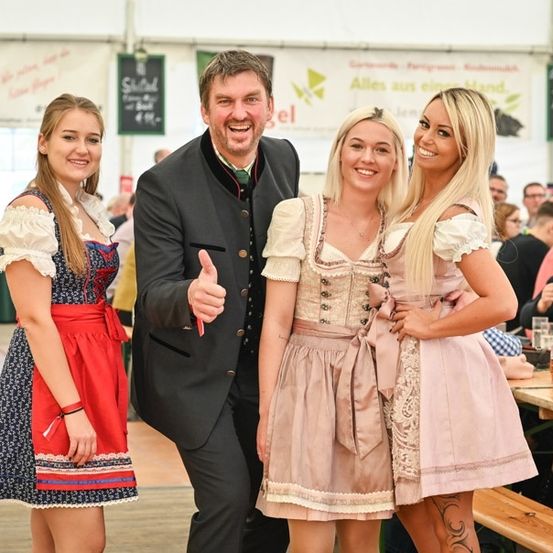 Drei Menschen in traditioneller deutscher Kleidung stehen bei einem Fest zusammen, lächeln und posieren für ein Foto. Ein Mann gibt einen Daumen hoch.