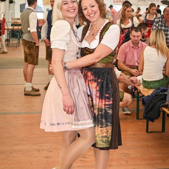 Zwei Frauen in traditioneller deutscher Kleidung lächeln und posieren für ein Foto auf einem Festival. Hinter ihnen sitzen und stehen Menschen, einige in traditioneller Kleidung.