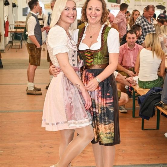 Zwei Frauen in traditioneller deutscher Kleidung stehen lächelnd in einer festlichen Umgebung, mit anderen Menschen im Hintergrund.
