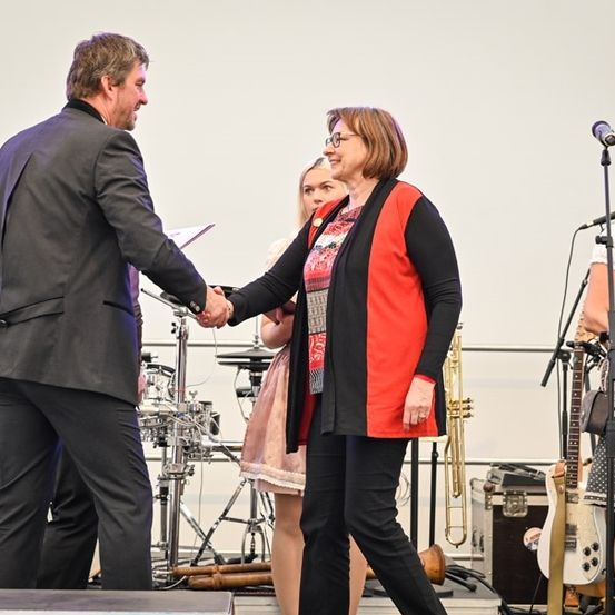 Ein Mann und eine Frau schütteln sich auf einer Bühne die Hände, mit Musikinstrumenten im Hintergrund.