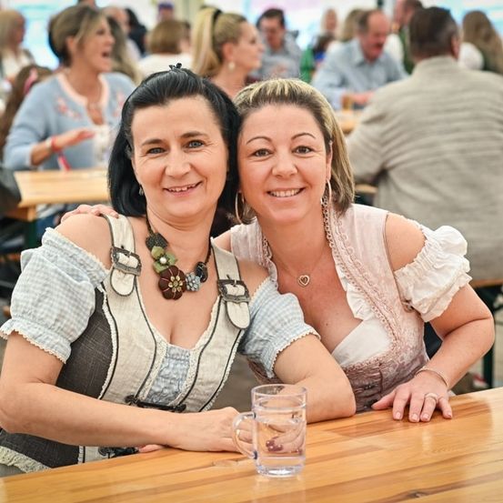 Zwei Frauen in traditioneller deutscher Kleidung lächeln und posieren für ein Foto bei einer überfüllten Outdoor-Veranstaltung. Sie sitzen an einem Holztisch mit Gläsern vor ihnen.