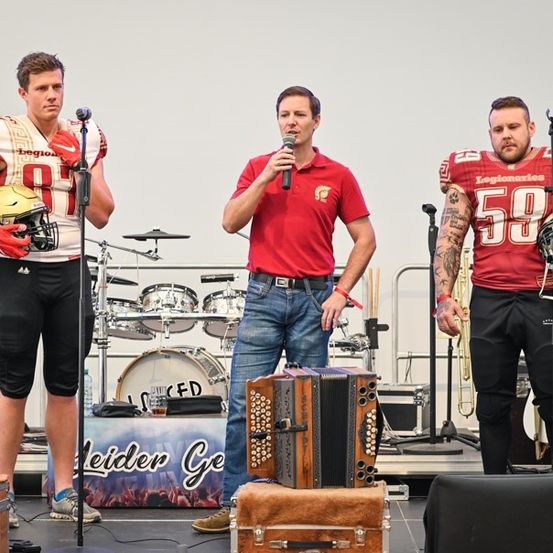 Drei Männer auf einer Bühne, einer hält ein Mikrofon, ein anderer spielt Akkordeon und der dritte spielt Trompete. Der Mann in der Mitte trägt ein rotes Hemd und Jeans. Hinter ihnen sind ein Schlagzeug und eine Gitarre zu sehen.