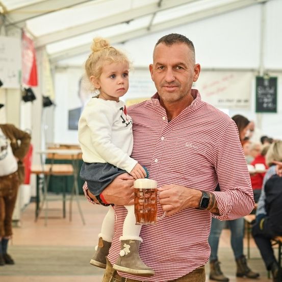 Ein Mann in einem rot-weiß karierten Hemd hält ein kleines Mädchen auf seiner Schulter und einen Bierkrug. Sie stehen in einem Zelt, in dem andere Leute an Tischen sitzen.