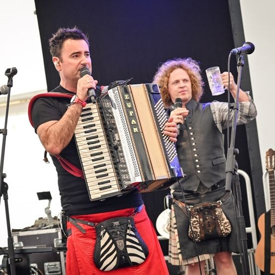 Ein Mann spielt Akkordeon und singt in ein Mikrofon, begleitet von einem anderen Mann mit einem Glas. Sie stehen auf einer Bühne mit Gitarren im Hintergrund.