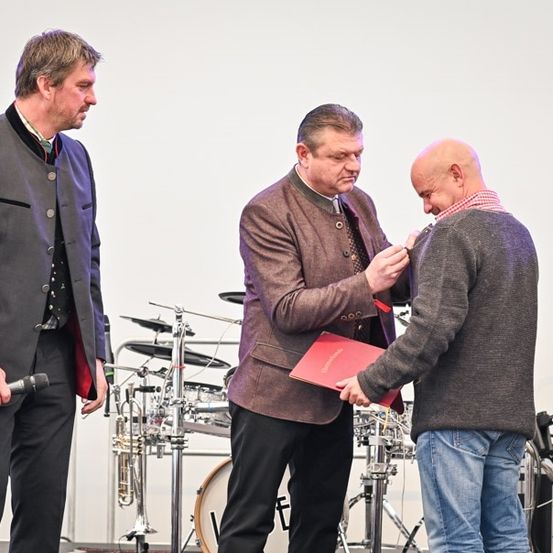 Drei Männer auf der Bühne, einer erhält von einem anderen eine Auszeichnung, während der dritte ein Mikrofon hält. Dahinter sind Musikinstrumente aufgebaut.