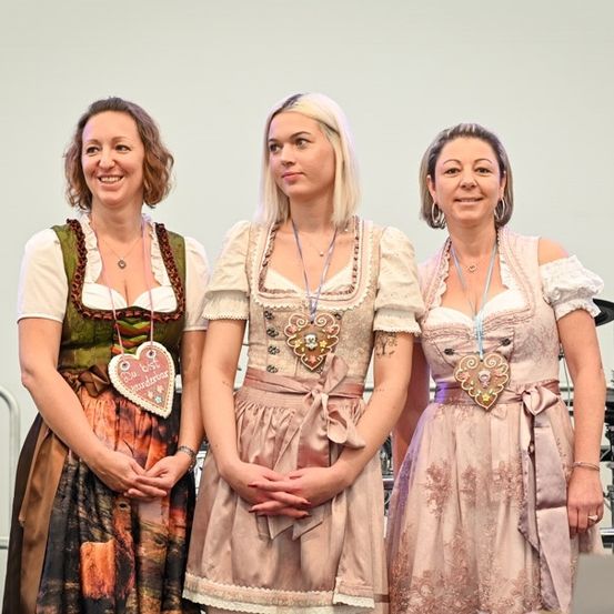 Drei Frauen in traditioneller deutscher Kleidung, lächelnd und zusammenstehend auf einer Bühne. Eine trägt ein grünes Kleid, eine andere ein rosa Kleid und die dritte ein weißes Kleid. Sie nehmen wahrscheinlich an einer kulturellen Veranstaltung teil.