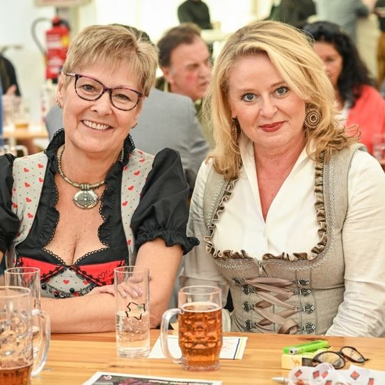 Zwei Frauen sitzen an einem Tisch mit Bierkrügen, eine in einem traditionellen deutschen Kleid und die andere in einer Bluse, beide lächeln.