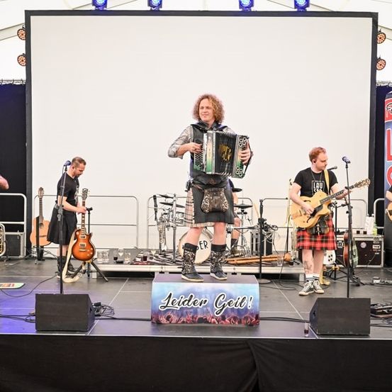 Eine Band tritt auf der Bühne auf, mit einer großen Leinwand im Hintergrund. Ein Mann spielt Akkordeon, ein anderer Gitarre, und zwei weitere spielen Schlagzeug und Bassgitarre. Die Bühne ist gut beleuchtet mit Scheinwerfern.